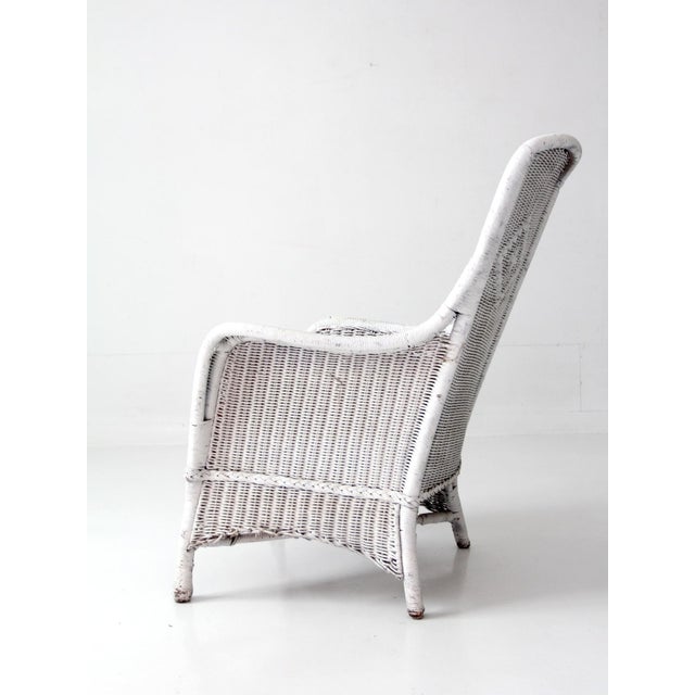 This antique white wicker porch chair features a tall sloped back, woven diamond detailing, and a softly aged painted...