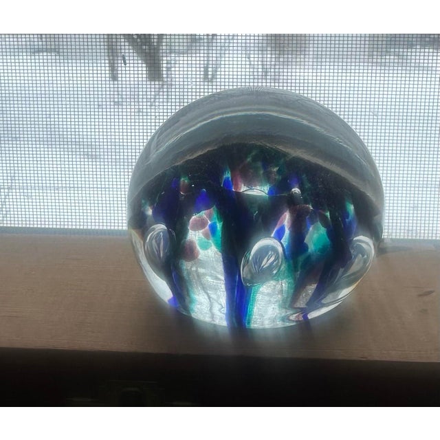A striking handblown art glass paperweight featuring a vibrant constellation of blue, teal, and mauve inclusions suspended...