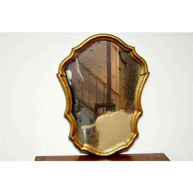 A small vintage Italian gilt wall mirror with an ornate gilded with a cartouche-shaped frame with scalloped edges. Maker’s...
