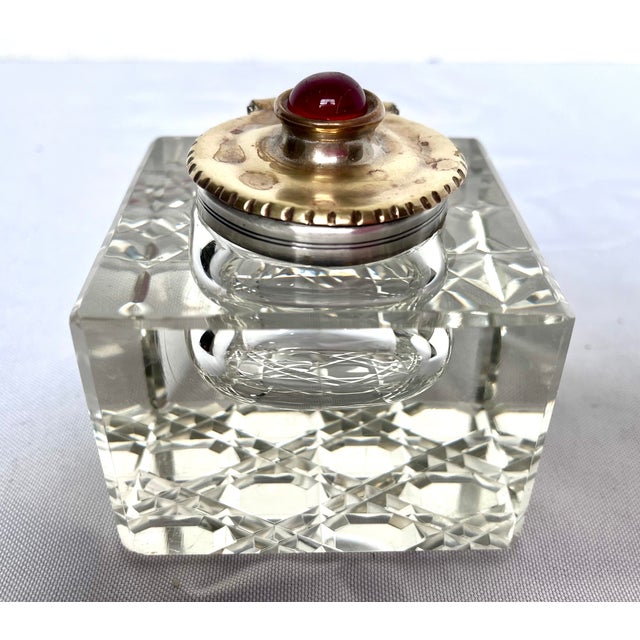 This antique inkwell features a square, cut crystal base paired with a silver and brass hinged lid.The lid is adorned with...