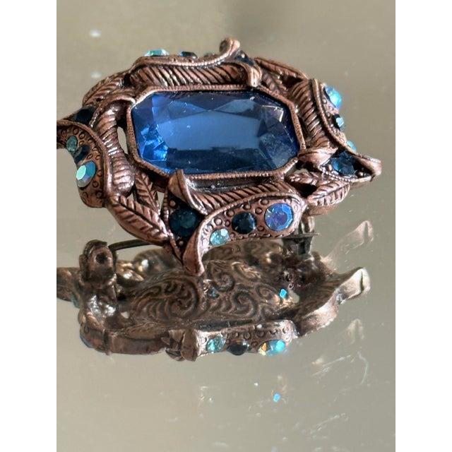 This 1970s Hollywood Regency brooch features a large emerald cut tanzanite-colored center stone framed by sculptural...