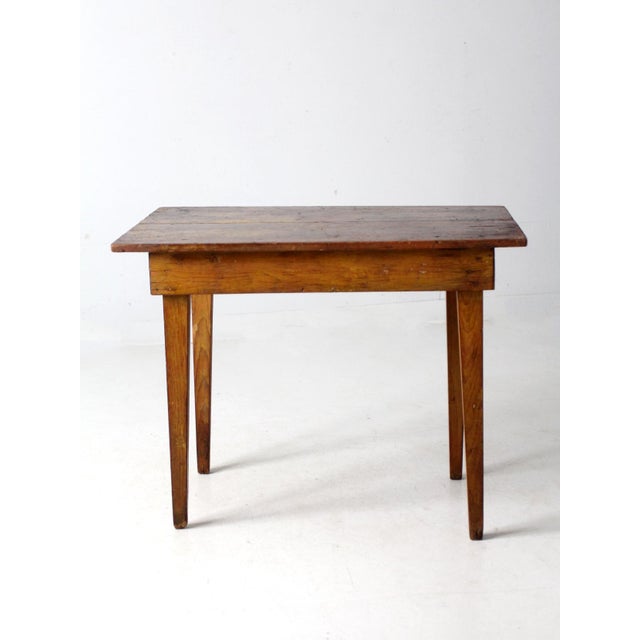 This antique primitive farmhouse table features a simple, rustic design with a top made of two planks and straight legs....