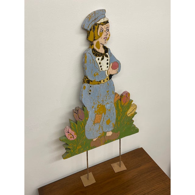 A charming vintage folk art painted wood figure depicting a stylized person standing among tulips, holding a rounded...