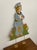 A charming vintage folk art painted wood figure depicting a stylized person standing among tulips, holding a rounded...