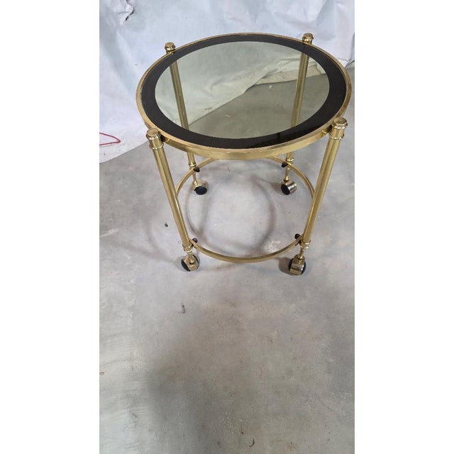 Round glass-top side table with a gold-tone metal frame, black border detail, and rolling casters. Measures 24″ tall × 20″...