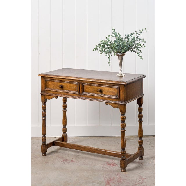 This oak side table features two paneled drawers with round wooden knobs for storage, while the beveled top showcases the...