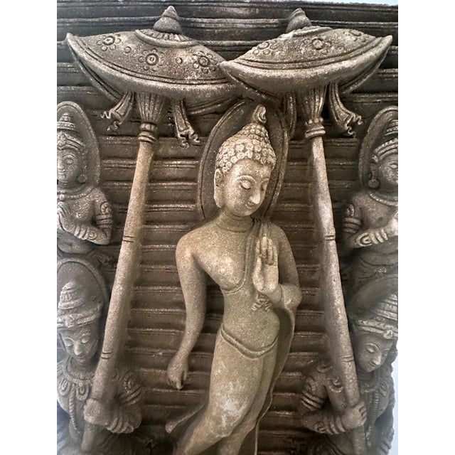 This intricately carved stone relief depicts a serene standing Buddha surrounded by attendants beneath ornate parasols, a...