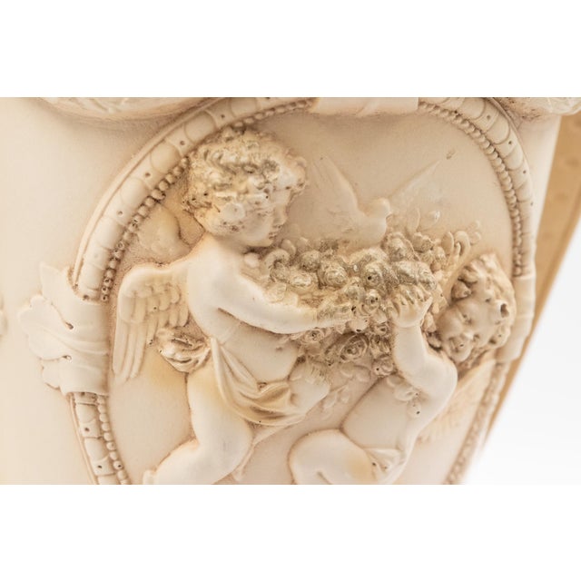 A large-scale Hydrastone chalkware urn in warm taupe, with twin handles and relief-molded cherub figures in the Greco-...