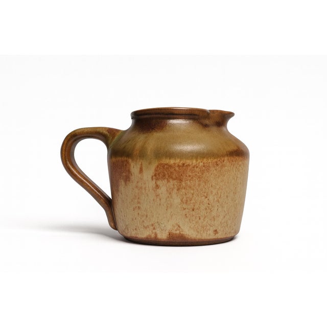 A bold studio pitcher finished in a copper orange glaze with brown undertones. The rounded body tapers toward the mouth,...