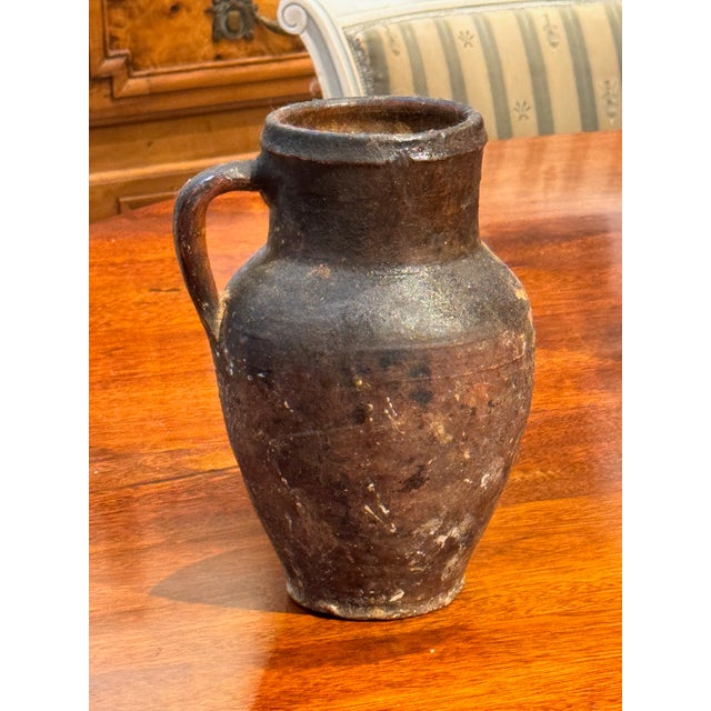 A small terra cotta jug with a single side handle and rounded body, showing an aged surface with darkened patina and wear...