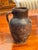 A small terra cotta jug with a single side handle and rounded body, showing an aged surface with darkened patina and wear...