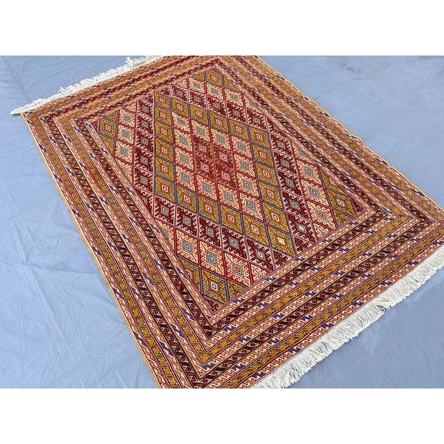 A flatwoven Mushwani kilim featuring an allover diamond lattice in warm gold, burgundy, ivory, and blue, framed by a...