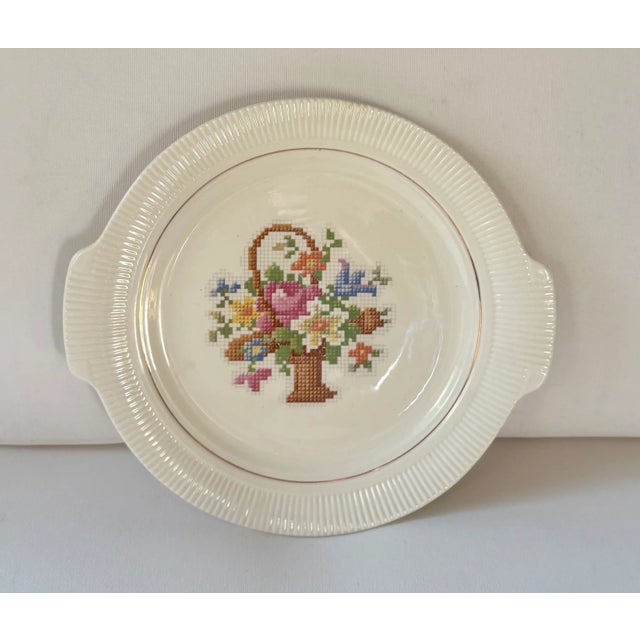 Artwork depicting a colorful needlepoint basket of flowers covers the face of ivory china cake plate produced by The Salem...