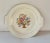 Artwork depicting a colorful needlepoint basket of flowers covers the face of ivory china cake plate produced by The Salem...