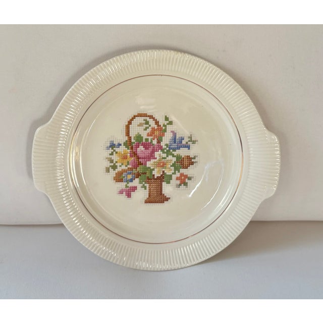 Artwork depicting a colorful needlepoint basket of flowers covers the face of ivory china cake plate produced by The Salem...