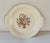 Artwork depicting a colorful needlepoint basket of flowers covers the face of ivory china cake plate produced by The Salem...