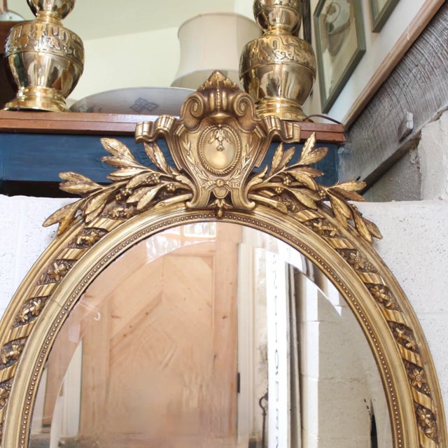 A boldly crested antique mirror with a ribboned cartouche and laurel branches, the gilded oval frame is composed of a...