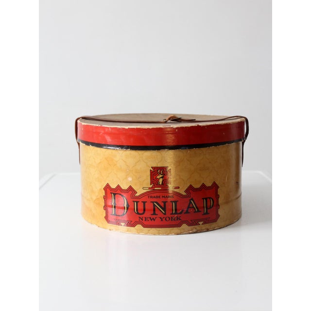 A vintage 1930s Dunlap New York hat box featuring bold red and black graphics against a warm gold patterned ground,...