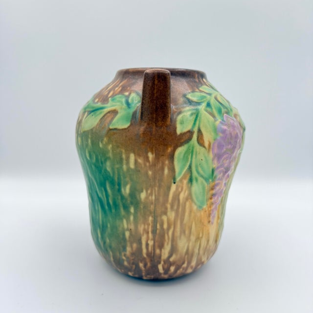 A richly glazed art pottery vase featuring dimensional wisteria blossoms and trailing leaves modeled in relief across the...