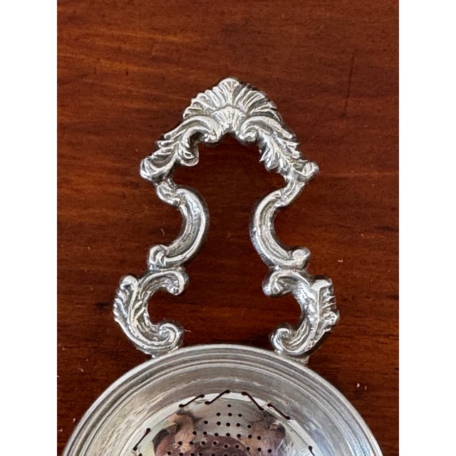 Elegant vintage silverplate tea strainer featuring a beautifully pierced bowl and ornate double scroll handles. The richly...