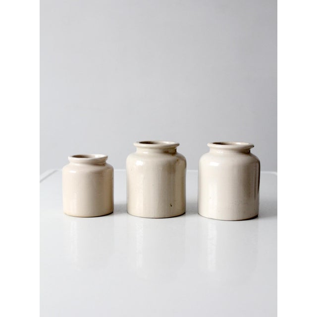 This set of three antique stoneware crocks dates to the early 20th century and features creamy ivory glaze, rounded...