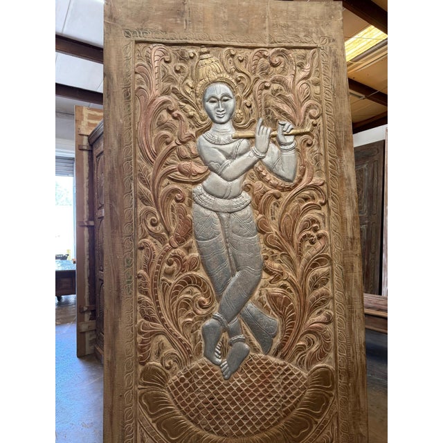 A striking hand-carved wooden wall panel featuring Lord Krishna in intricate relief, surrounded by ornate floral and vine...