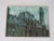 Mid Century painting captures a city building scene and is painted on burlap instead of a traditional canvas, with a pink...
