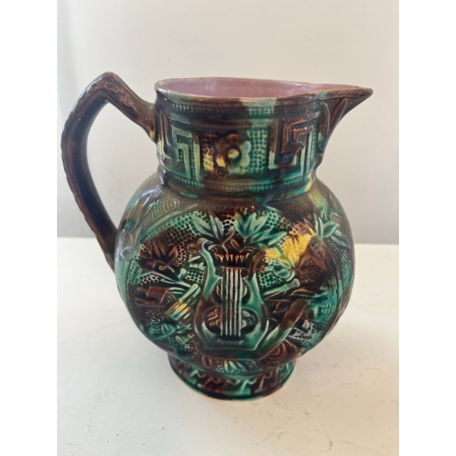 Two ceramic majolica pitchers. One in chocolate, browns greens and yellows with a harp decoration on either side the other...