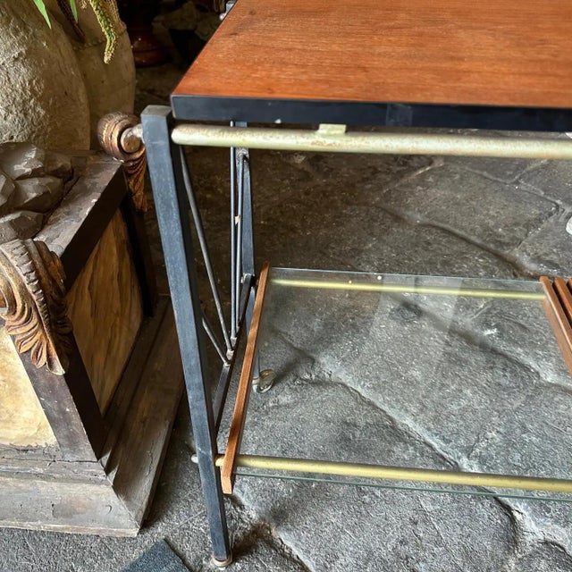 This cart has a black metal frame with a minimalist, geometric design that is typical of the Mid-Century Modern style. The...