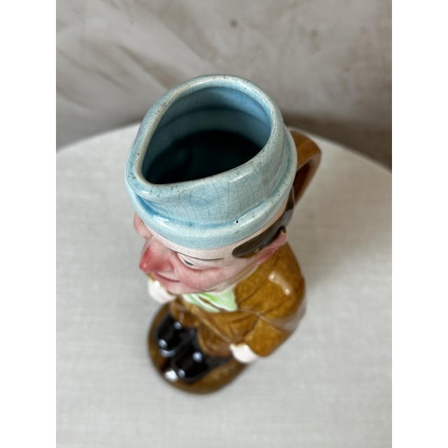 A charming, humorous earthenware pitcher made in the early 20th century by the Saint-Amand-les-Eaux factory. It depicts a...