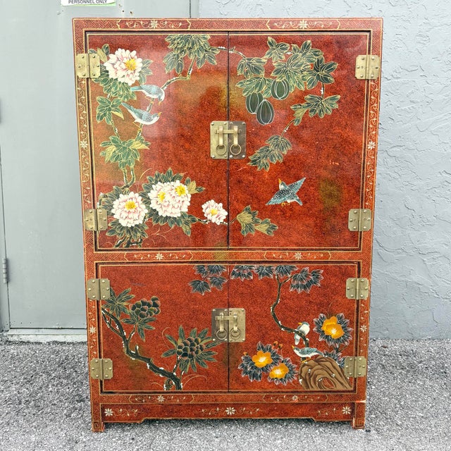 A striking vintage Chinese armoire featuring a rich red lacquer-style finish with elaborate hand-painted botanical and...