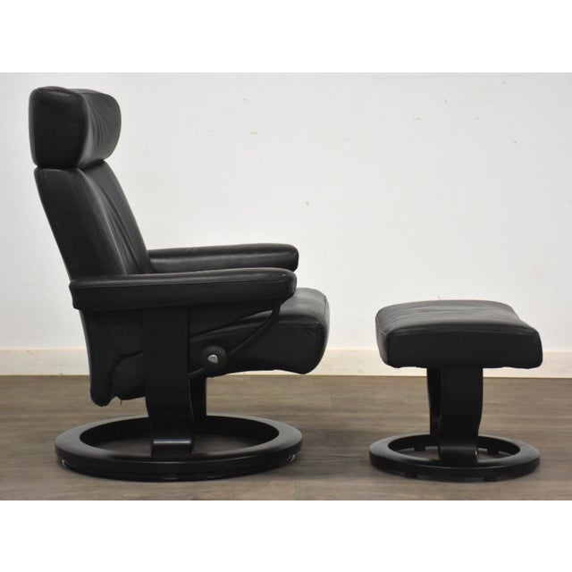 A modern black leather reclining lounge chair and ottoman made by Ekornes Stressless. Seat height is 16”. Ottoman is...