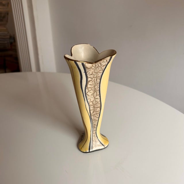 A striking small ceramic vase featuring a sculptural flared form and warm yellow tone with hand-painted blue accents. The...