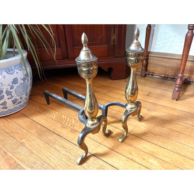 Pair of midcentury brass andirons in the Federal style, topped with elegant finials and crafted with brass. These firedogs...