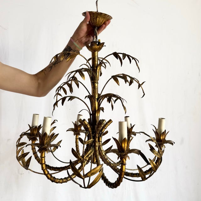 This striking Hollywood Regency chandelier features sculptural faux bamboo arms adorned with delicate gilt leaves, forming...