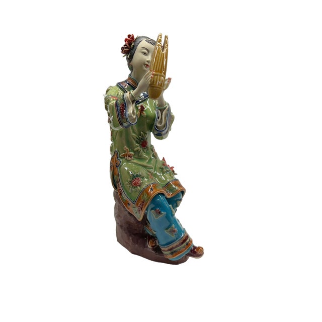 This exquisite porcelain figurine captures the serene beauty and artistic tradition of a young woman playing a sheng, a...