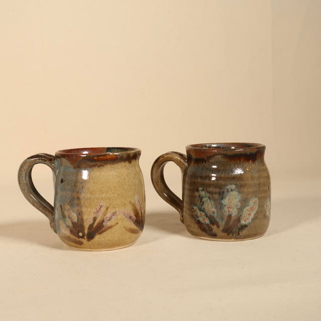 This charming pair of handmade studio pottery mugs showcases warm earth-toned glazes, subtle painterly floral accents, and...
