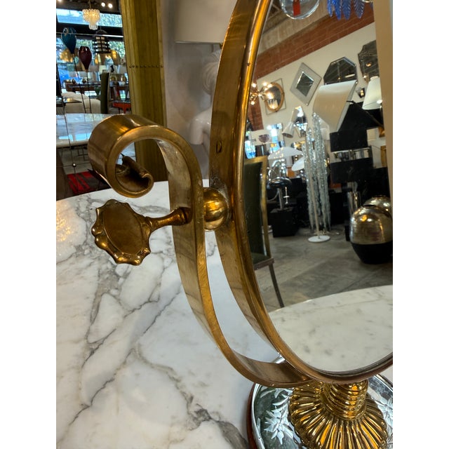 Charming vintage vanity mirror featuring a classic round brass frame with elegant curved supports and a decorative...