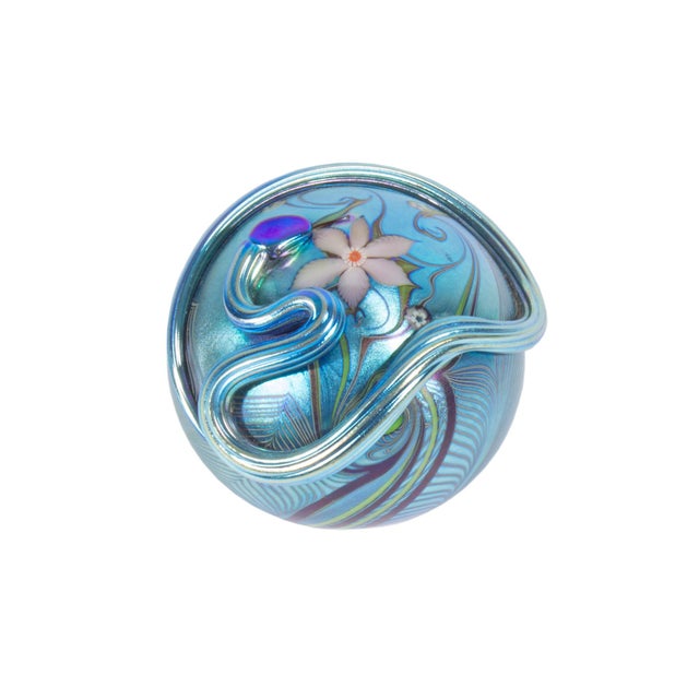 A 1979 iridescent art glass paperweight by Orient and Flume. This round paperweight features an intricate floral design...