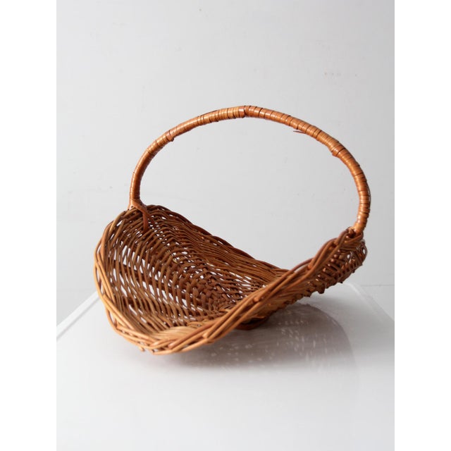 A sculptural vintage wicker kindling basket featuring a dramatic crescent shape and fixed woven handle. The elevated base...