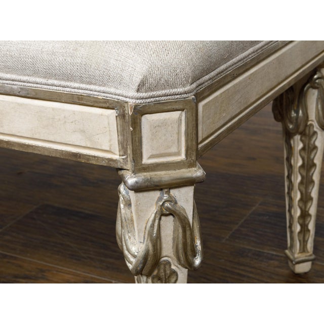 An Italian Midcentury painted bench with silver gilt carved foliage motifs, tapered legs and custom linen upholstery. This...