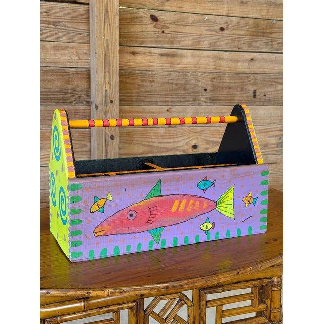 A cheerful vintage wooden caddy featuring whimsical hand-painted fish, colorful geometric accents, and a striped carrying...