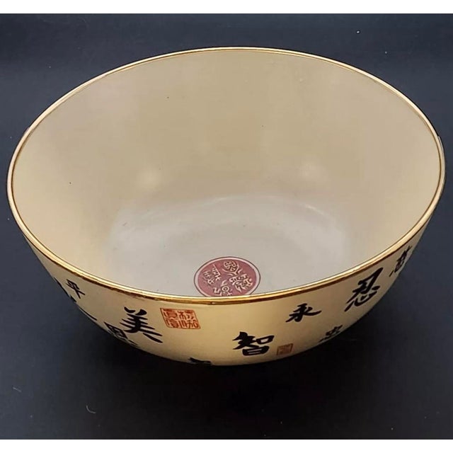 Large deep Chinese bowl in cream with black and red Chinese writing and symbols. The bowl has a metallic gold gilt rim and...