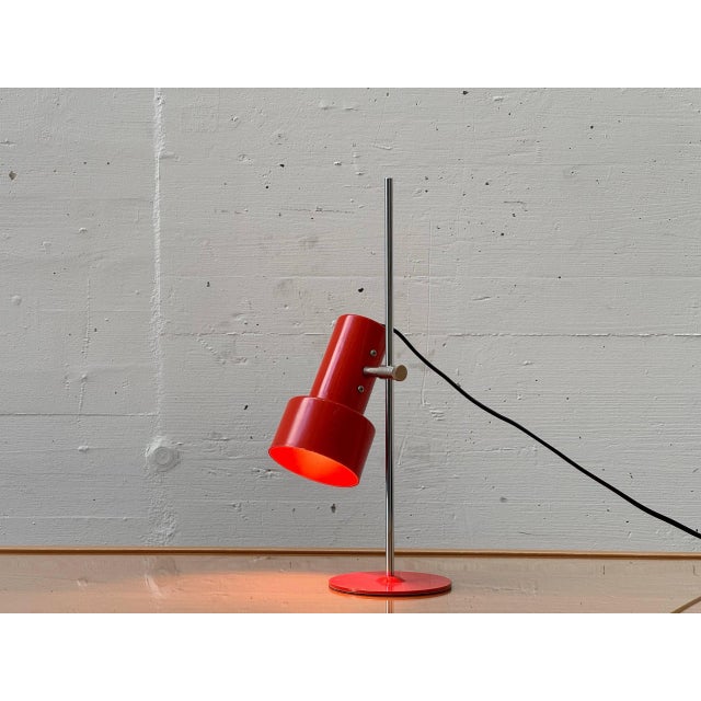 A distinctive table lamp in a functional mid-century design with a clear, technical aesthetic. The cylindrical lamp head,...