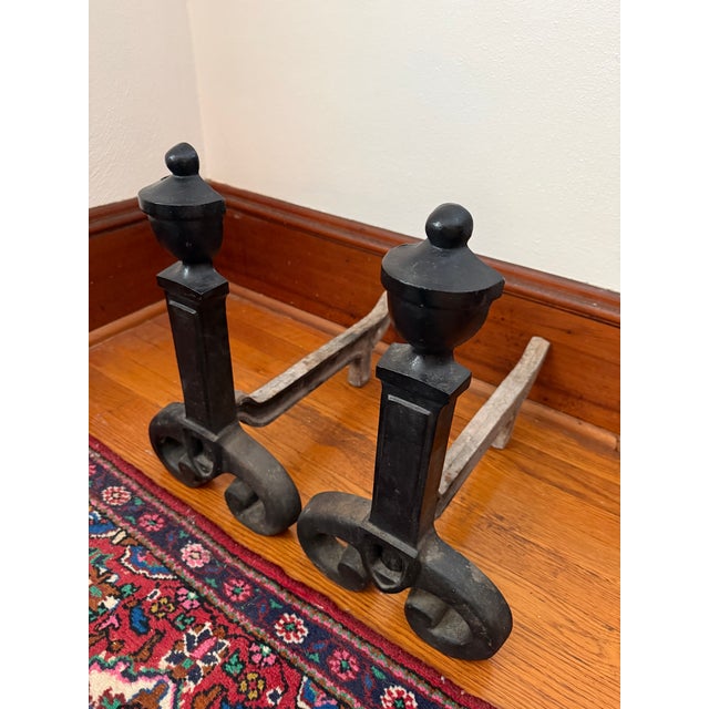 Pair of black wrought iron andirons. Each one has an urn finial on a square tapered post. Large scrolled feet sit at the...