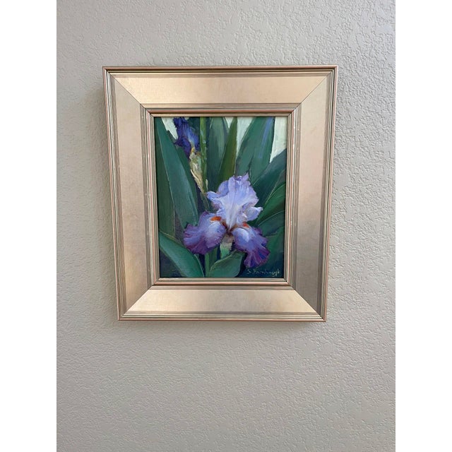 This oil painting captures the delicate beauty of an iris, its soft lavender petals contrasting with the lush green leaves...