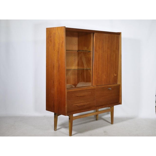 Teak highboard with a glass display case, sliding door, and delicate base. The glass display case with two glass shelves...