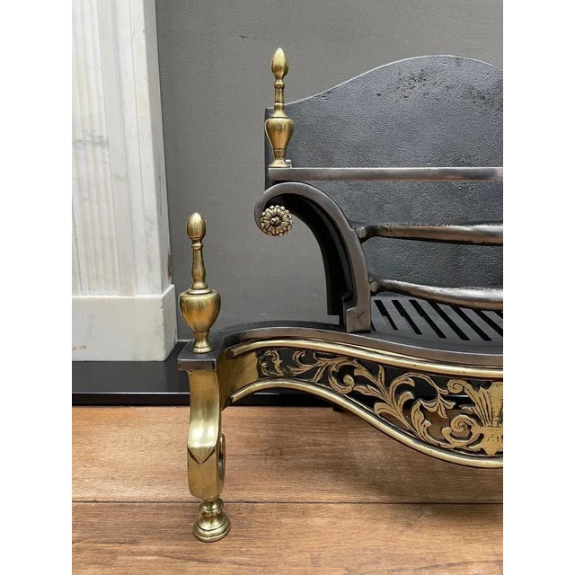 An English brass and wrought iron fire grate. The scrolled and shaped legs surmounted by finials, flanking a curved and...