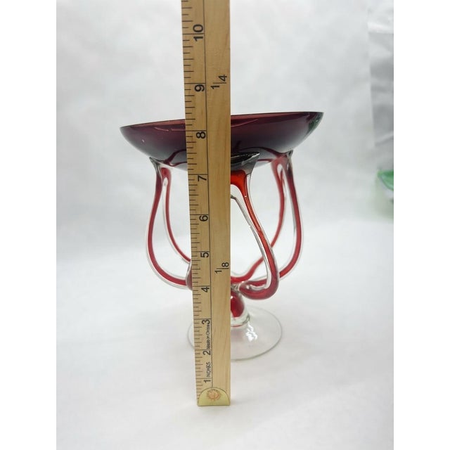 Striking mid-century modern art glass compote featuring a deep cranberry-to-ruby red bowl cradled by an elaborate,...