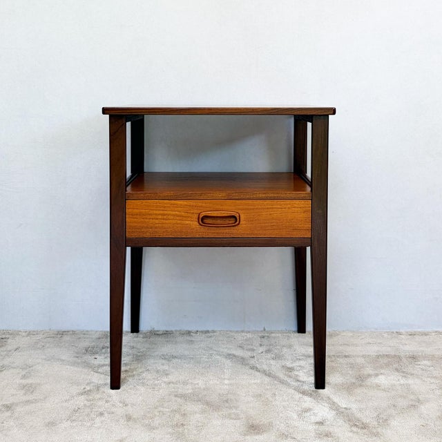 Constructed with teak veneer top and drawer front over a frame structure. Four tapered legs in darker stained wood support...